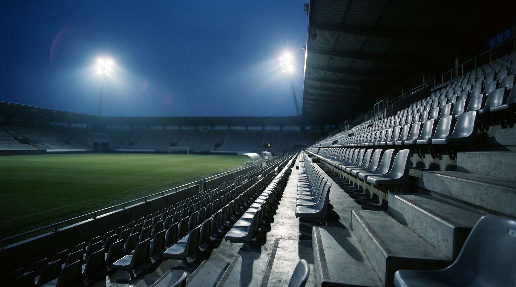 Flutlicht geht in der Dämmerung an, Blick aus der leeren Tribüne über das Spielfeld